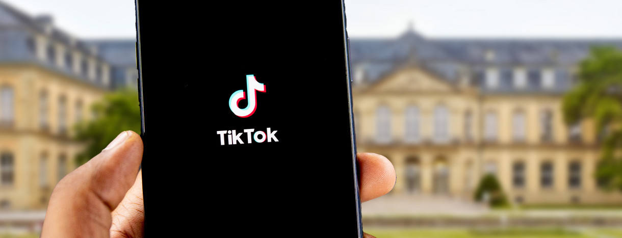 Hand mit Smartphone in der Hand, auf dem Bildschirm sieht man die App TikTok, im Hintergrund ist das Neue Schloss Stuttgart zu sehen