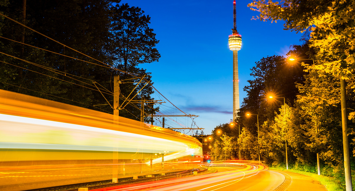 Vorbeifahrender Zug mit Lichtspuren vor dem beleuchteten Fernsehturm Stuttgart in der Dämmerung.