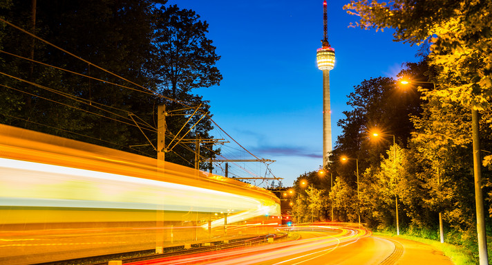 Vorbeifahrender Zug mit Lichtspuren vor dem beleuchteten Fernsehturm Stuttgart in der Dämmerung.