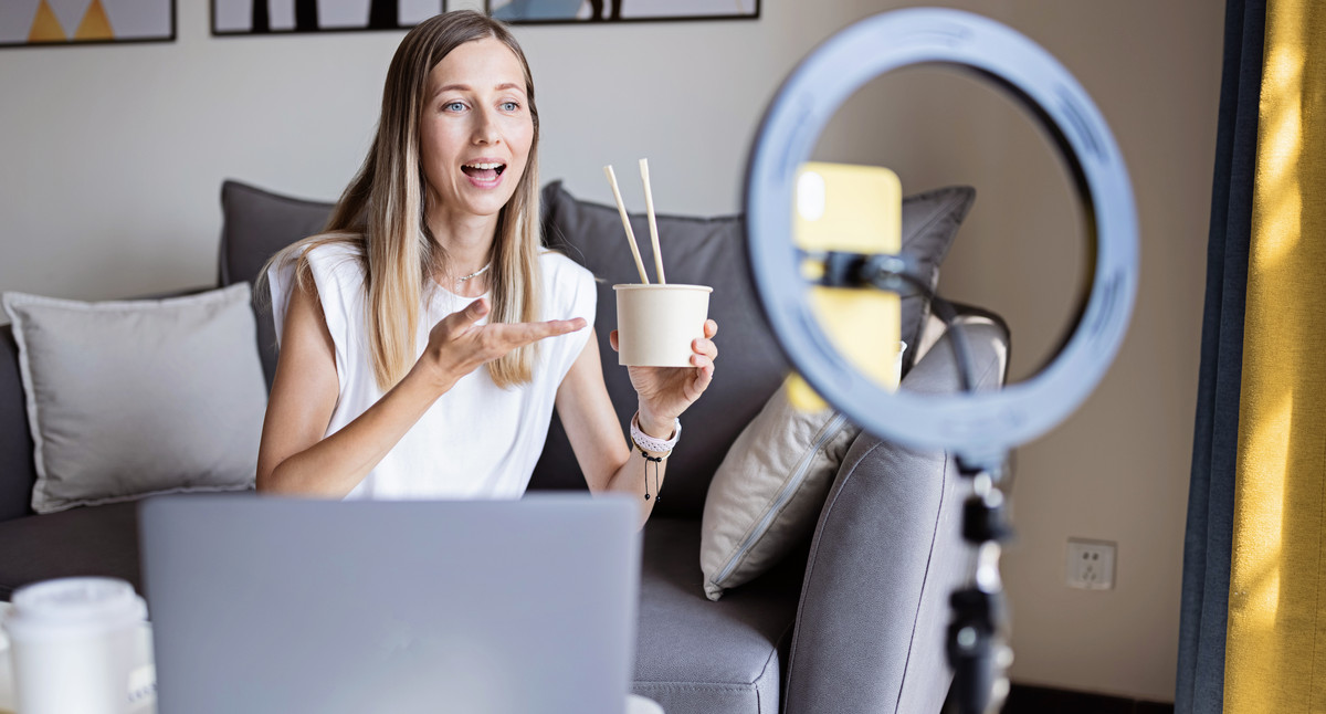 Junge Frau erstellt mit ihrem Smartphone und dem Licht einer Ringlampe Foto- oder Videoinhalte für Social Media. Als Influencerin streamt sie live und kommuniziert mit ihren Followerinnen und Followern in sozialen Netzwerken.