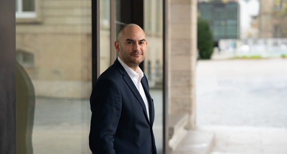 Finanzminister Danyal Bayaz steht vor dem Kunstgebäude am Schlossplatz in Stuttgart, im Hintergrund sind der Landtag und weitere Gebäudestrukturen zu sehen.