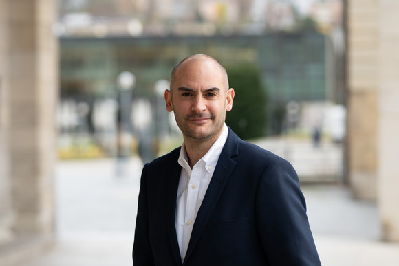 Finanzminister Danyal Bayaz steht am Schlossplatz in Stuttgart, im Hintergrund ist moderne Architektur sowie ein Teil des Platzes zu sehen.