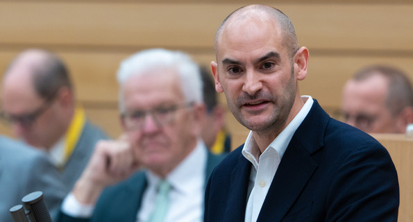 Finanzminister Danyal Bayaz spricht im Plenarsaal des Landtags am Redepult, im Vordergrund scharf zu sehen. Im unscharfen Hintergrund sitzt Ministerpräsident Winfried Kretschmann.