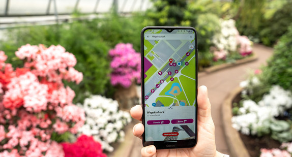 Smartphone mit der neuen Wilhelma App im Bildschirm, im Hintergrund ein Gewächshaus der Wilhelma