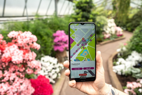 Smartphone mit der neuen Wilhelma App im Bildschirm, im Hintergrund ein Gewächshaus der Wilhelma