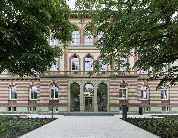 Frontalansicht des historischen Backsteingebäudes des MINT-Exzellenzgymnasiums in Bad Saulgau. Der verglaste Eingangsbereich wird von großen Bäumen eingerahmt.