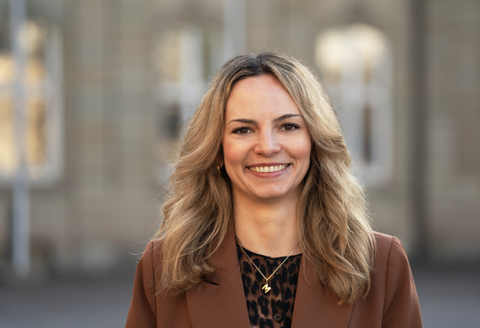 Porträt einer lächelnden Frau mit schulterlangen, leicht gewellten blonden Haaren. Sie trägt einen braunen Blazer über einem gemusterten Oberteil sowie eine feine Halskette und blickt direkt in die Kamera. Im Hintergrund ist ein unscharfes Gebäude zu sehen.