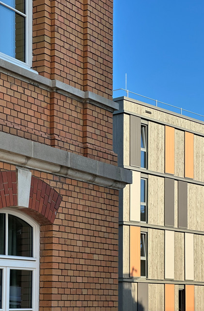 Detailansicht zweier Gebäudefassaden. Links die historische Backsteinfassade mit Rundbogenfenstern, rechts der moderne Neubau mit Holzverkleidung und farbigen Fassadenelementen.