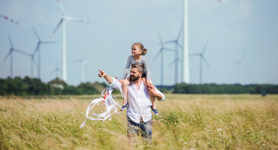 Ein junger Vater läuft mit seiner Tochter, die auf seinen Schultern sitzt, über eine Wiese. Im Hintergrund sind Windräder zu sehen. / Foto: stock.adobe.com / Halfpoint 