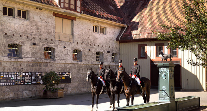 Drei in traditioneller Uniform gekleidete Reiter zu Pferd, die vor historischen Gebäuden des Haupt- und Landgestüts Marbach stehen, mit Kopf- und Mauerreliefs im Hintergrund sowie einem Brunnen mit Pferdeskulptur im Vordergrund.