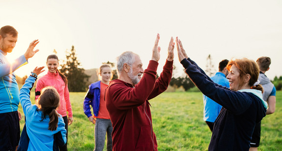 Mehrere Menschen stehen in Sportoutfits auf einer Wiese, zwei Personen stehen sich gegenüber und klatschen sich gegenseitig ab. Foto: stock.adobe.com/Halfpoint 