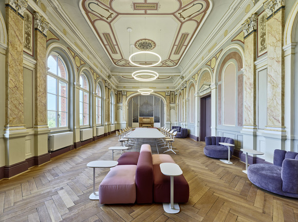 Lang gestreckter, historisch verzierter Raum mit hohen Rundbogenfenstern, Parkettboden und einer Orgel am Ende des Saals. Moderne Sitzmöbel und Tische bilden verschiedene Arbeits- und Aufenthaltsbereiche.