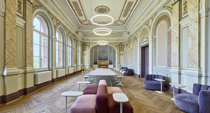 Lang gestreckter, historisch verzierter Raum mit hohen Rundbogenfenstern, Parkettboden und einer Orgel am Ende des Saals. Moderne Sitzmöbel und Tische bilden verschiedene Arbeits- und Aufenthaltsbereiche.