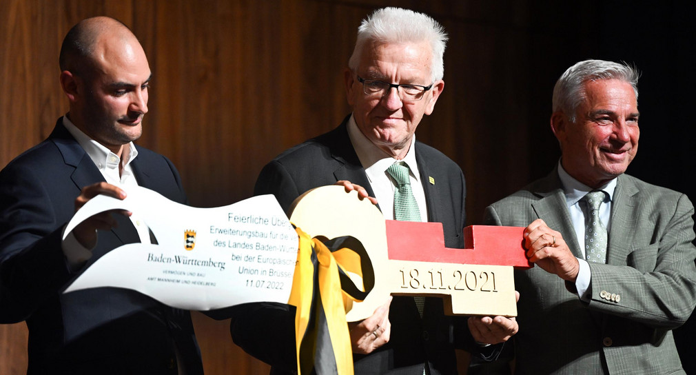 von links nach rechts: Finanzminister Danyal Bayaz, Ministerpräsident Winfried Kretschmann und Innenminister Thomas Strobl halten in der Landesvertretung in Brüssel einen symbolischen Schlüssel in den Händen.