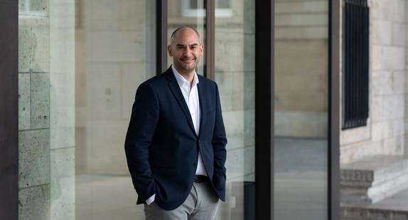 Finanzminister Danyal Bayaz steht vor dem Kunstgebäude am Schlossplatz in Stuttgart, angelehnt an eine Glasfassade, die Details der Gebäudestruktur widerspiegelt.