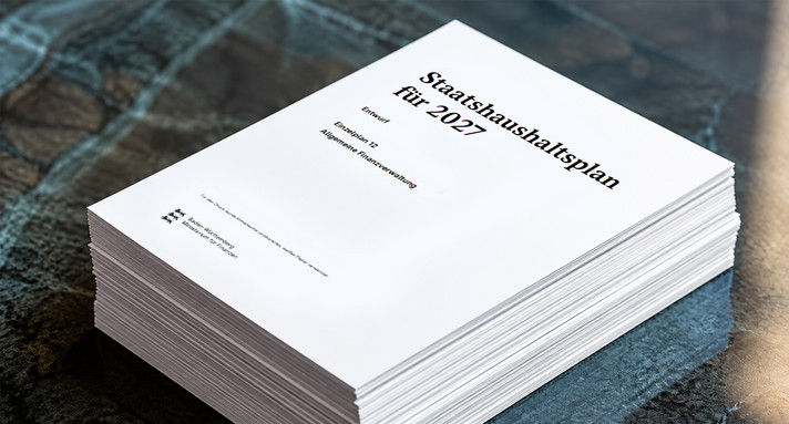 Ein Stapel gedruckter Haushaltspläne liegt auf einer dunklen Tischplatte. Auf dem obersten Deckblatt steht „Staatshaushaltsplan für 2027, Entwurf, Einzelplan 12, Allgemeine Finanzverwaltung“.