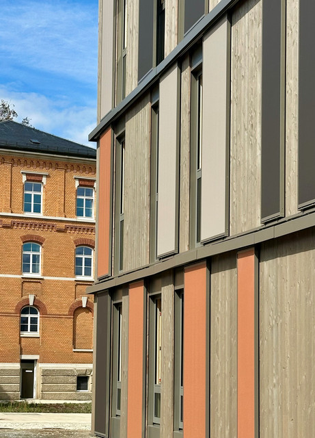 Blick entlang der modernen Holzfassade mit grauen und terrakottafarbenen Elementen. Im Hintergrund ist das historische Backsteingebäude mit großen Fenstern zu sehen.
