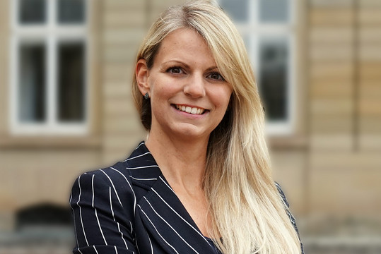 Porträt einer lächelnden blonden Frau vor einem unscharfen Gebäudehintergrund. Sie trägt ein dunkelblaues, fein gestreiftes Sakko und blickt direkt in die Kamera.