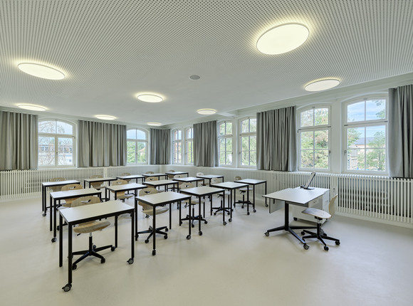 Heller Klassenraum mit großen Fenstern, grauen Vorhängen und weißen Wänden. Einzelne Tische mit schwarzen Gestellen und Stühle auf Rollen sind im Raum verteilt.