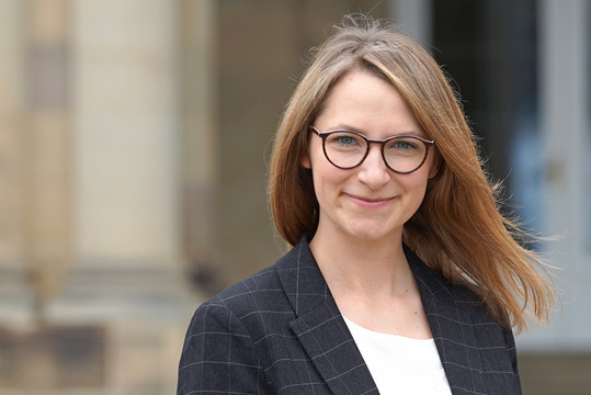 Porträt einer lächelnden Frau mit braunen, schulterlangen Haaren und runder Brille. Sie trägt ein dunkles kariertes Sakko über einer weißen Bluse und steht vor einem unscharfen Gebäudehintergrund.