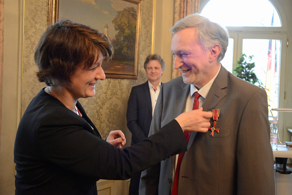 Staatssekretärin Gisela Splett steckt Dankwart von Loeper das Bundesverdienstkreuz ans Jacket. Im Hintergrund: Frank Mentrup, Oberbürgermeister der Stadt Karlsruhe.