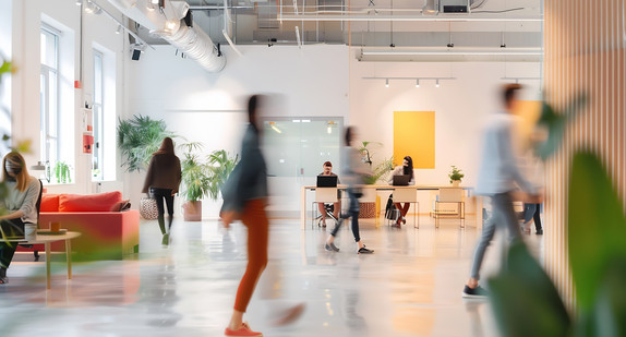 Moderner Coworking Space mit offenen Arbeitsbereichen, Schreibtischen, Pflanzen und mehreren Personen in Bewegung.